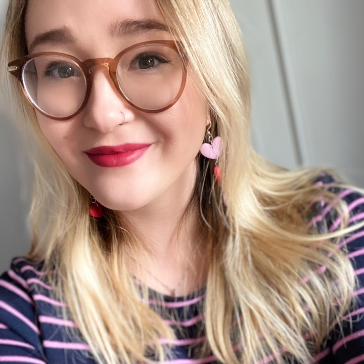 Liv, smiling, with blonde hair, round glasses, red lipstick and pink heart earrings